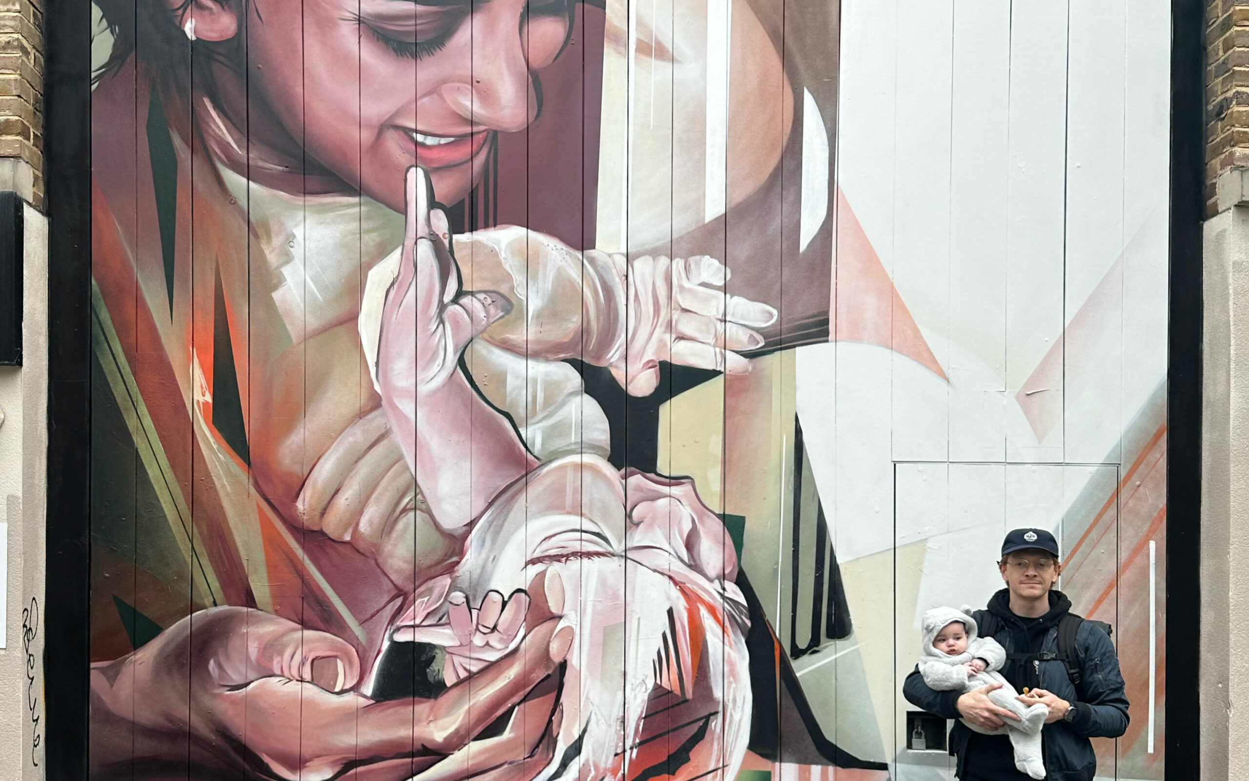 Artist Jake Attewell stands holding a baby in front of one of his large murals depicting a parent cradling a child in a blend of realism and abstract forms.