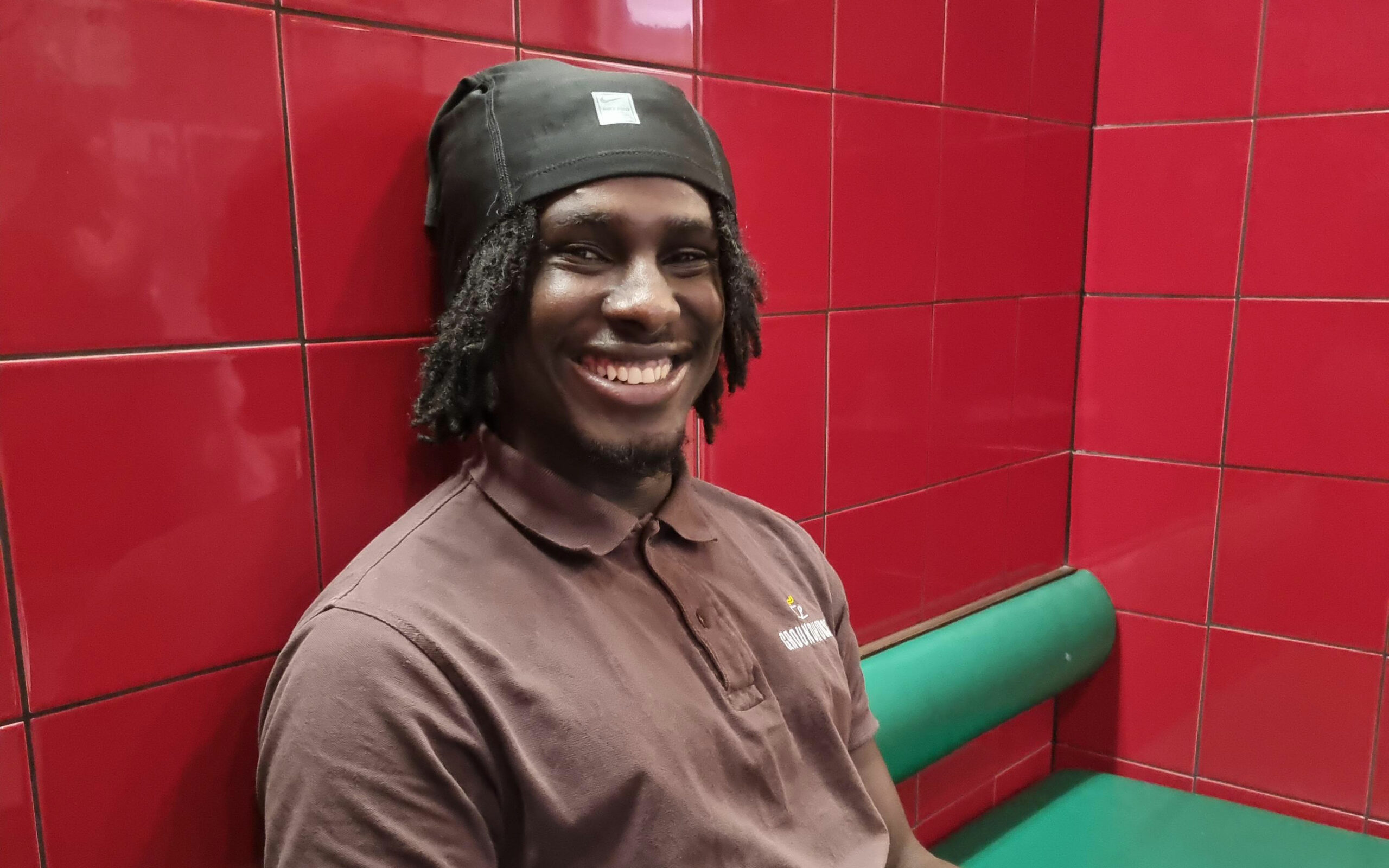Mustafa, a café team member, sits smiling in his Groundhouse uniform against a red tiled wall.