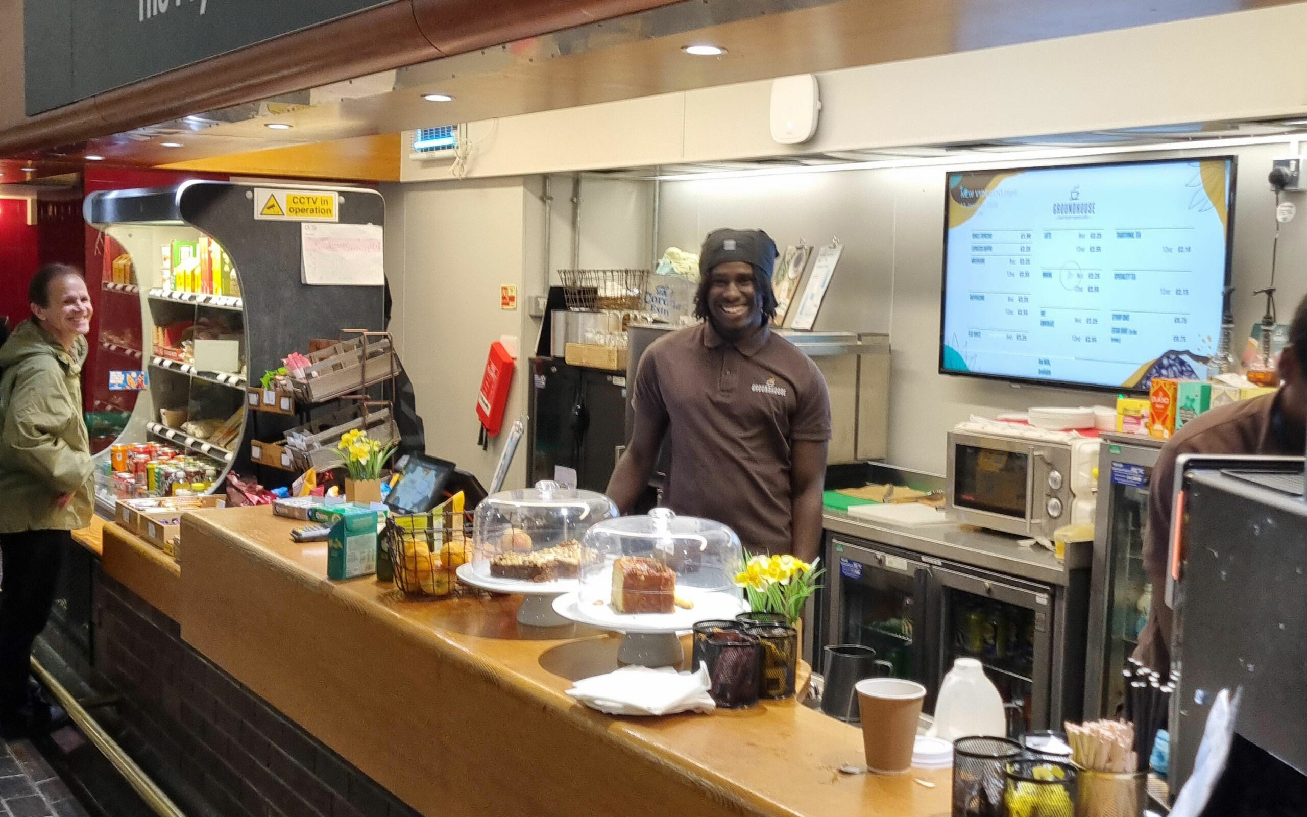 Mustafa smiles behind the café counter with cakes, fruit and drinks displayed in front of him.