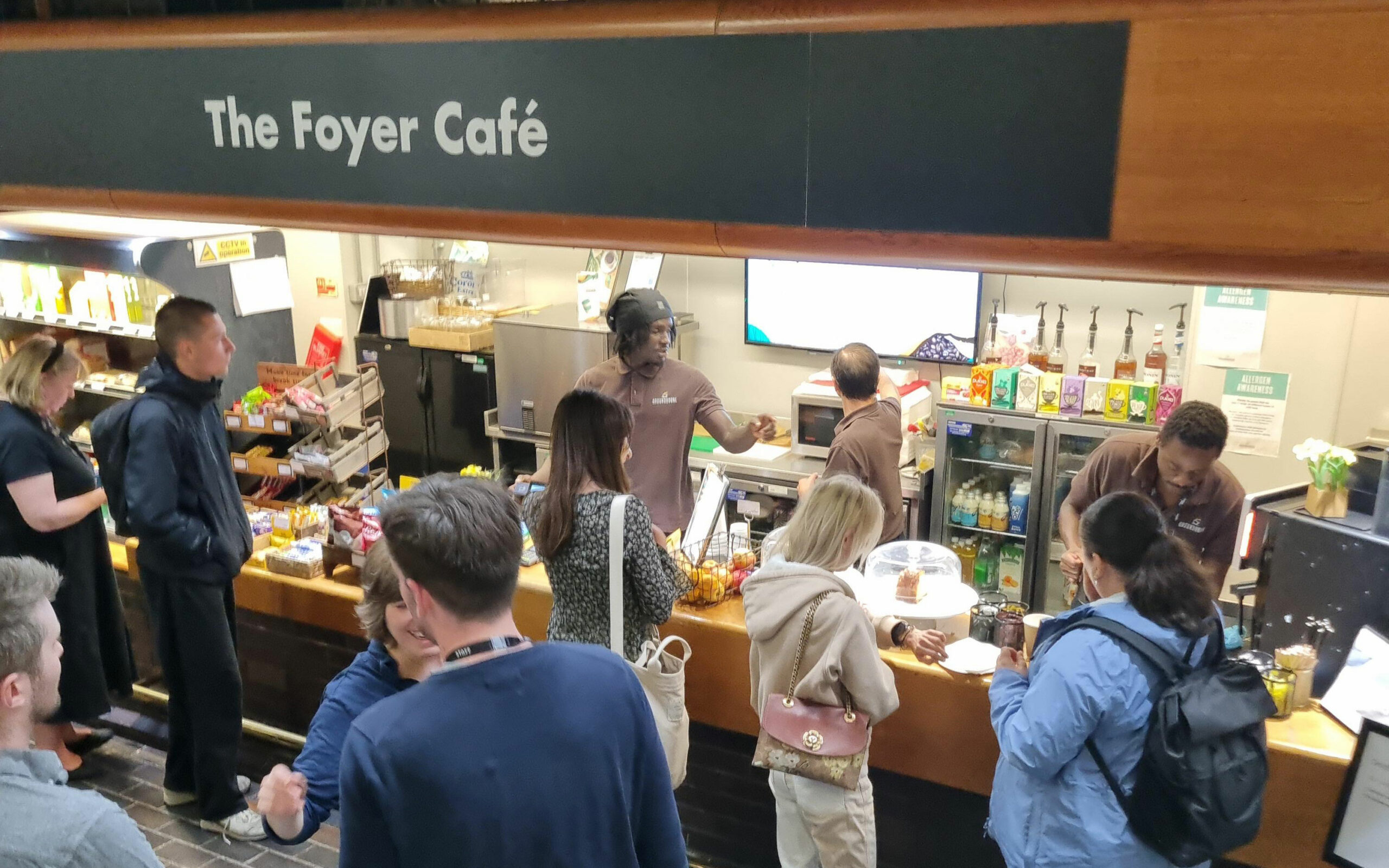 Staff at The Foyer Café serve customers at a busy counter with drinks, snacks and cakes on display.
